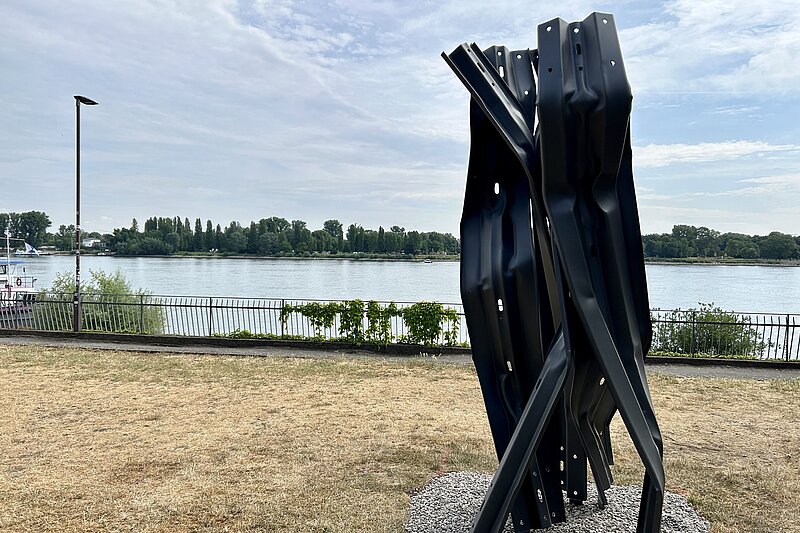 Eine Skulptur aus Leitplanken steht hochkant auf der Wiese am Rheinufer. Im Hintergrund fließt der Rhein.