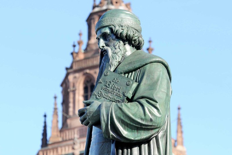 Vor blauem Himmel steht eine Statue von Gutenberg. Im Hintergrund ist der Dom zu sehen.