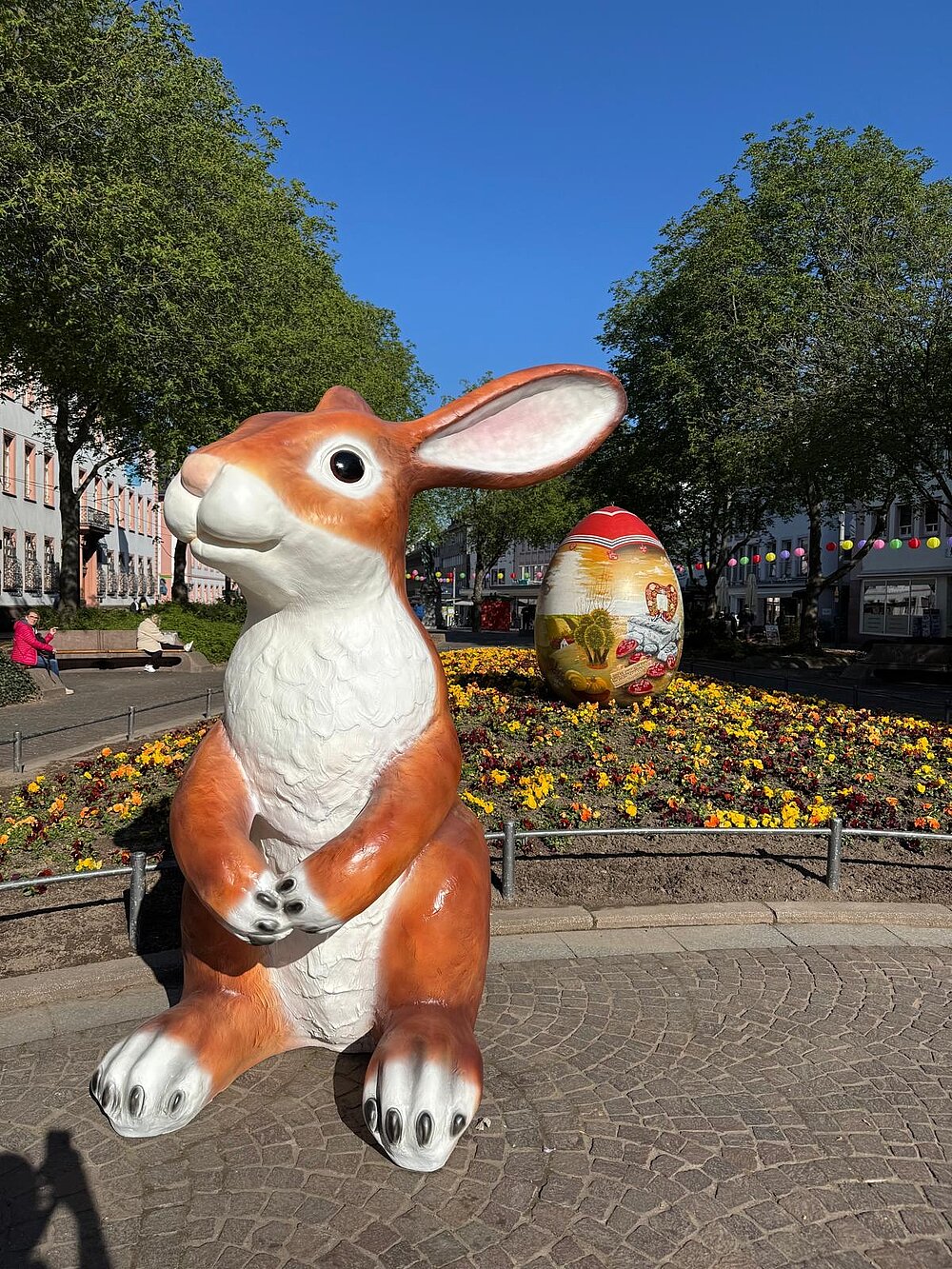 Auf dem Schillerplatz steht eine große Osterhasenfigur, schräg dahinter ist ein farbenfrohes, großes Osterei zu sehen.