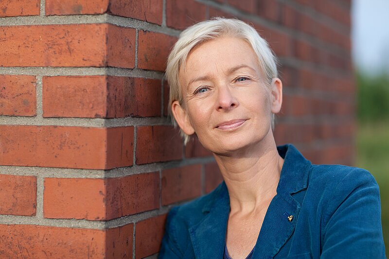 Eine blonde Frau in blauem Blazer lehnt an einer roten Backsteinwand. Die Frau auf dem Bild ist die Mainzer Stadtschreiberin Dörte Hansen.