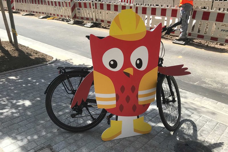 Ein Fahrrad steh am Wegesrand. Daran lehnt das Symbol der Sozialen Stadt: Eine rote Eule, die eine gelbe Warnweste, einen gelben Helm und gelbe Schuhe trägt.