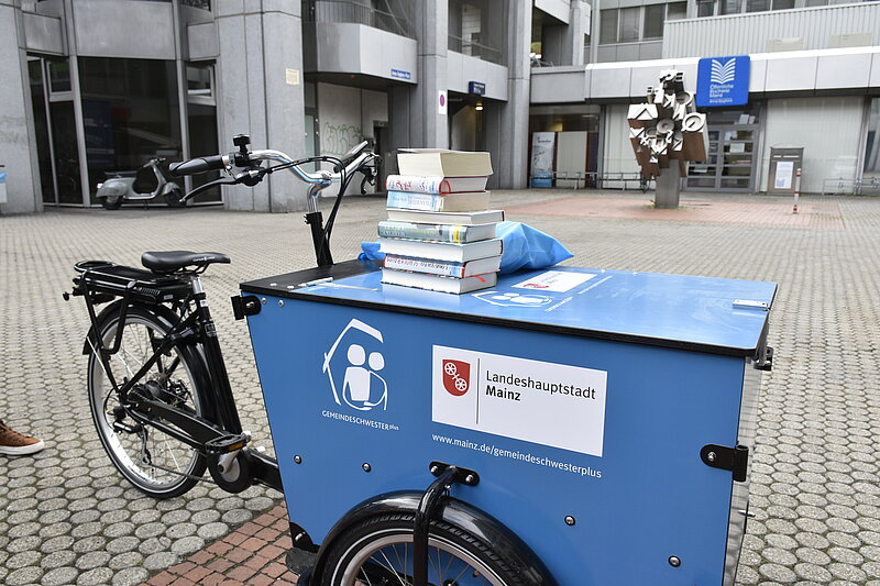 Ein blaues Lastenrad vor der Bibliothek Anna Seghers. Auf dem Lastenrad sind die Logos der Landeshauptstadt Mainz und der Gemeindeschwester plus zu sehen. Ein Bücherstapel liegt auf der Beförderungskiste des Lastenrades.