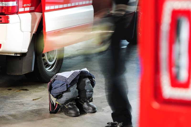 Ein kleiner Ausschnitt eines Feuerwehrautos ist abegebildet. Davor steht jemand und wartet, man sieht nur die Rückseite bis zur Hüfte, sie ist verwischt. Vor der Person liegt eine Feuerwehrhose, die noch in Schuhe steckt.. 