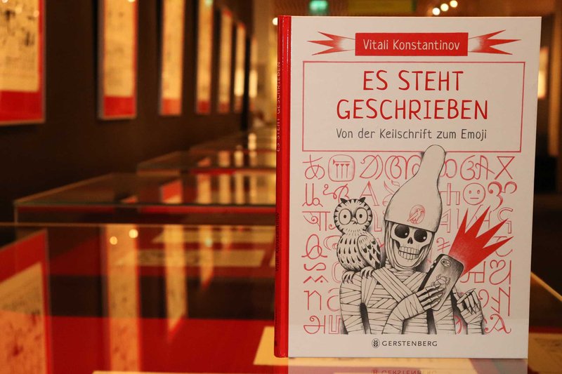 Das Buch "Es steht geschrieben - Von der Keilschrift zum Emoji" von Vitali Konstantinov ist zu sehen. Im Hintergrund sind unschrf Ausstellungsexponate zu erkennen.