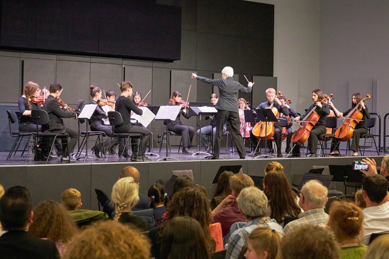 Zu sehen ist ein Jugendorchester mit Dirigent auf der Bühne. Im Vordergrund Publikum.