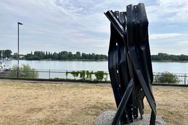 Eine Skulptur aus Leitplanken steht hochkant auf der Wiese am Rheinufer. Im Hintergrund fließt der Rhein.